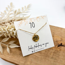 Hand-stamped 70th birthday gold coin necklace with a satellite beaded chain displayed on a white jewelry card that reads "70 looks fabulous on you." The necklace rests on a wooden tray with dried white botanical floral props in the background. Handcrafted milestone birthday gift for women by Jenica Jewelry.