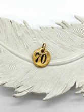 Hand-stamped gold pebble charm with the number 70, displayed on a textured white feather ceramic dish. SEO-optimized image for custom milestone 70th birthday gifts, personalized number pendants, and customizable charm necklaces by Jenica Jewelry.