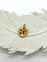 Hand-stamped gold pebble charm with the number 70, displayed on a textured white feather ceramic dish. SEO-optimized image for custom milestone 70th birthday gifts, personalized number pendants, and customizable charm necklaces by Jenica Jewelry.