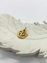 Hand-stamped gold coin charm with the number 70, displayed on a textured white feather dish. Rustic personalized pendant for custom milestone birthday jewelry, lucky number necklaces, or charm bracelets by Jenica Jewelry.