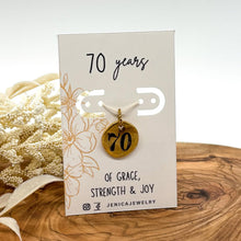 Handcrafted 70th birthday gold pebble charm mounted on a white display card reading "70 years OF GRACE, STRENGTH & JOY" with subtle floral line art. The round gold pendant is hand-stamped with the number "70" in bold black ink, sitting upright on a natural wood surface with dried white botanicals in the background.