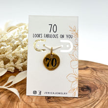 A small round gold pendant stamped with the number "70" mounted on a white jewelry display card. The card reads "70 LOOKS FABULOUS ON YOU" with delicate floral line artwork and the handle @JENICAJEWELRY at the bottom. The jewelry card rests on a warm wood slice alongside dried white botanical sprigs against a clean white backdrop.