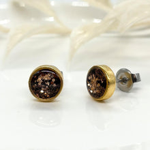 Pair of handmade round stud earrings in gold-plated bezels filled with shimmering brown glitter sealed in clear resin. The left earring faces forward to display the sparkling inlay, while the right earring shows the side profile, surgical steel post, and earring back. White botanical accents blur softly in the background.