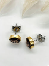 Angled top-down view of handmade gold stud earrings featuring round resin bezels with bronze glitter inlays. Both earrings display the hypoallergenic stainless steel posts and metallic butterfly clutch backs resting on a clean white surface with soft white botanical accents above.