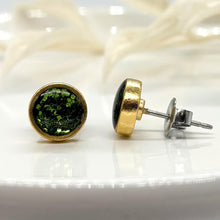 Pair of round stud earrings set in gold-plated bezels resting on a white ceramic display plate with blurred white botanicals behind. The left earring faces forward, showing its sparkling Christmas green glitter resin inlay. The right earring is turned to display its side profile, gold bezel rim, hypoallergenic stainless steel post, and butterfly clutch back.