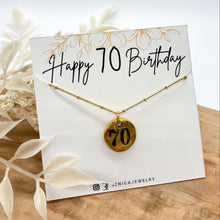 Gold hand-stamped pebble charm necklace displayed on a white branded jewelry card reading "Happy 70 Birthday" in cursive script with floral line art. The organic round gold pendant features the number "70" stamped in black ink on a delicate beaded chain. Resting on a light wood surface with dried white botanicals framing the display card.