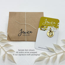 Packaging display showcasing eco-friendly kraft paper gift wrapping tied with white string alongside a branded Jenica Jewelry display card holding a sample gold number charm. Overlay text at the bottom reads "Sample item shown. All orders arrive wrapped in our signature kraft packaging!" Framed with delicate white dried botanicals against a clean background.