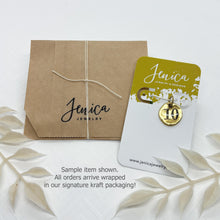 Packaging display showcasing eco-friendly kraft paper gift wrapping tied with white string alongside a branded Jenica Jewelry display card holding a sample gold number charm. Overlay text at the bottom reads "Sample item shown. All orders arrive wrapped in our signature kraft packaging!" Framed with delicate white dried botanicals against a clean background.