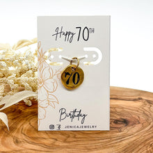 Handcrafted 70th birthday gold pebble charm mounted on a white display card reading "Happy 70TH Birthday" in modern script with delicate floral line art accents. The round gold-plated charm is hand-stamped with the number "70" in bold black ink, sitting upright on a warm wooden board next to soft white dried botanicals.