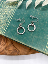 A pair of small silver dangle hoop earrings resting on a teal vintage book decorated with ornate gold filigree. Each earring features a hammered circular ring charm suspended from a small connector loop on French wire ear hooks with clear rubber stoppers. The book sits on a warm wood surface with a white leafy botanical branch overhead.