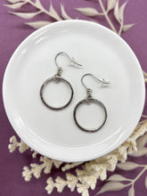 A pair of silver dangle hoop earrings displayed on a small white ceramic dish. Each earring features a small textured gold ring suspended from French hook ear wires with clear rubber backs. The dish rests on a purple background accented with dried white botanical stems.