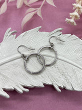 A pair of silver dangle hoop earrings resting on a white feather-shaped ceramic tray. Each earring features a circular silver ring suspended from a smaller textured connector ring on French wire hooks with clear rubber backs. The tray is set against a textured pink background styled with soft white botanical accents.