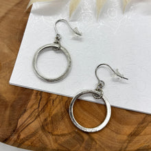 A pair of silver dangle hoop earrings presented on a white card with a subtle floral emboss pattern. Each earring consists of a hammered, circular gold hoop suspended from a small textured gold ring connected to French ear wires with clear rubber stoppers. The card rests on a natural wood surface with a white leaf accent above.