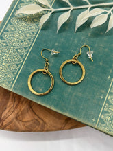 A pair of gold dangle hoop earrings resting on a teal vintage book with ornate gold filigree details. Each earring features a circular gold hoop suspended from a textured gold jump ring on French wire hooks with clear rubber backings. The book is set on a wooden surface with a white leaf sprig overhead.