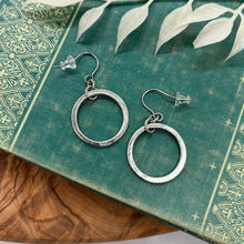 A pair of silver dangle hoop earrings resting on a teal vintage book with ornate gold filigree details. Each earring features a circular gold hoop suspended from a textured gold jump ring on French wire hooks with clear rubber backings. The book is set on a wooden surface with a white leaf sprig overhead.