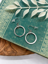 A pair of silver dangle hoop earrings resting on a teal vintage book with ornate gold filigree details. Each earring features a circular gold hoop suspended from a textured gold jump ring on French wire hooks with clear rubber backings. The book is set on a wooden surface with a white leaf sprig overhead.