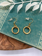 A pair of small gold dangle hoop earrings resting on a teal vintage book decorated with ornate gold filigree. Each earring features a hammered circular ring charm suspended from a small connector loop on French wire ear hooks with clear rubber stoppers. The book sits on a warm wood surface with a white leafy botanical branch overhead.