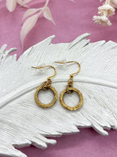 A pair of small gold circle dangle earrings rests on a decorative white feather ceramic tray set against a soft pink background with dried white botanicals. Each earring features a textured round hoop pendant suspended from gold French hook ear wires fitted with clear rubber backings.