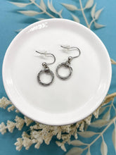 A pair of small silver circle dangle earrings displayed on a small white ceramic dish set against a vibrant turquoise background with dried white botanicals. Each earring features a textured round hoop suspended from silver French ear hooks with clear rubber stoppers.