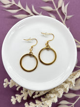 A pair of gold dangle hoop earrings displayed on a small white ceramic dish. Each earring features a small textured gold ring suspended from a French hook ear wires with clear rubber backs. The dish rests on a purple background accented with dried white botanical stems.
