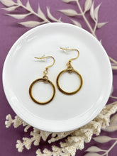A pair of gold dangle hoop earrings displayed on a small white ceramic dish. Each earring features a small textured gold ring suspended from a French hook ear wires with clear rubber backs. The dish rests on a purple background accented with dried white botanical stems.