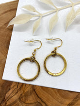 A pair of gold dangle hoop earrings presented on a white card with a subtle floral emboss pattern. Each earring consists of a hammered, circular gold hoop suspended from a small textured gold ring connected to French ear wires with clear rubber stoppers. The card rests on a natural wood surface with a white leaf accent above.