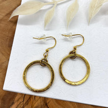 A pair of gold dangle hoop earrings presented on a white card with a subtle floral emboss pattern. Each earring consists of a hammered, circular gold hoop suspended from a small textured gold ring connected to French ear wires with clear rubber stoppers. The card rests on a natural wood surface with a white leaf accent above.