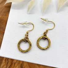 A pair of gold dangle hoop earrings placed on a white floral-patterned display card. Each earring features a small, rustic gold ring charm suspended from a textured connector loop attached to French wire ear hooks with clear rubber stoppers. The card is set against a wooden background with a white leafy branch overhead