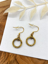 A pair of gold dangle hoop earrings placed on a white floral-patterned display card. Each earring features a small, rustic gold ring charm suspended from a textured connector loop attached to French wire ear hooks with clear rubber stoppers. The card is set against a wooden background with a white leafy branch overhead