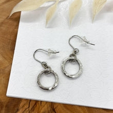 A pair of small silver circle dangle hoop earrings resting on a white floral-embossed card over a natural wood surface with dried white botanical foliage. Each earring features a textured round hoop suspended from silver French hook ear wires with clear rubber backings.