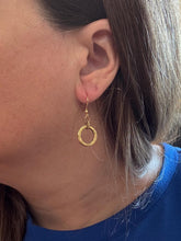 A side-profile photo modeling a single small gold circle dangle earring. The earring features a textured round hoop pendant dangling from a delicate gold French wire ear hook, shown against a woman with straight brown hair wearing a bright blue top.